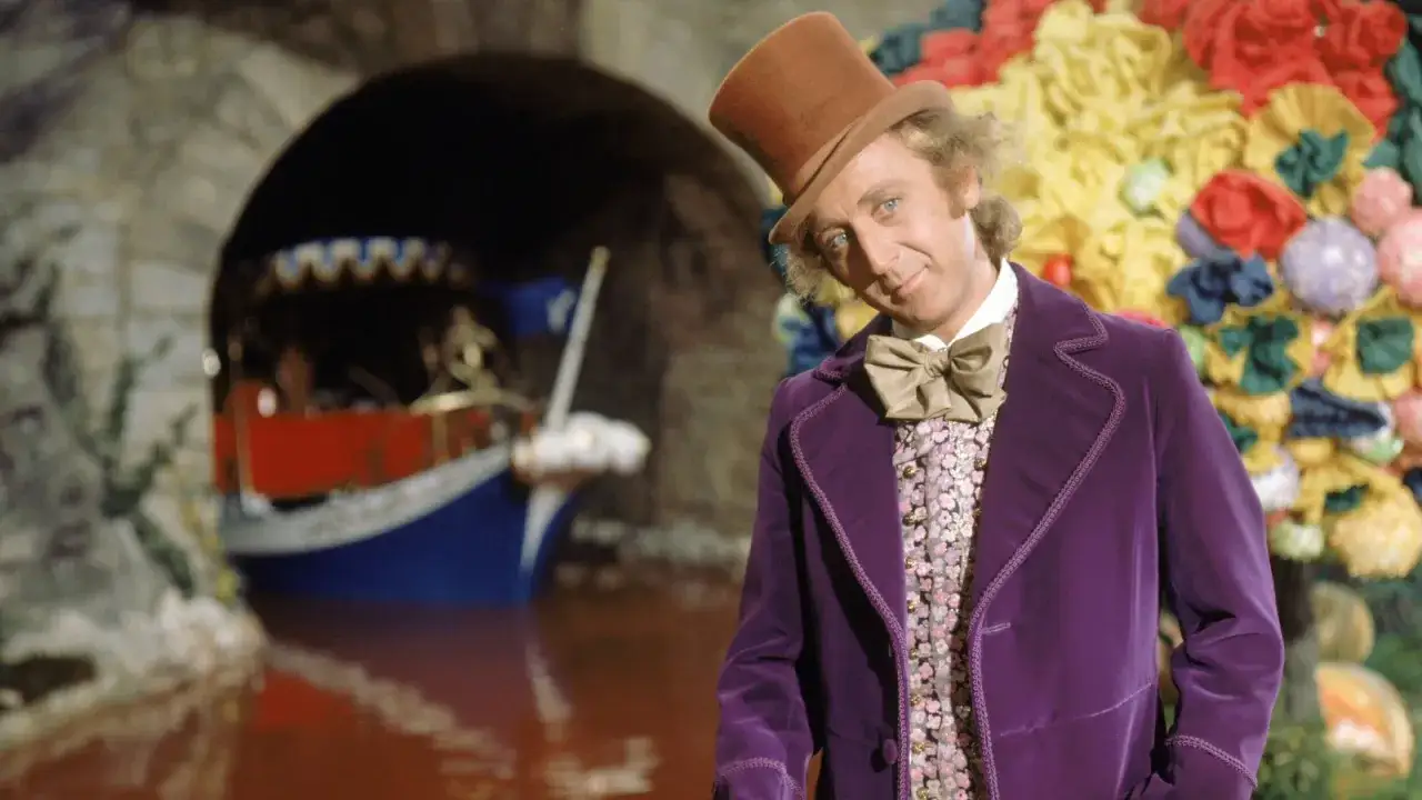 Gene Wilder, i sin ikoniska lila sammetsrock och hatt, står vid en chokladflod med en båt.