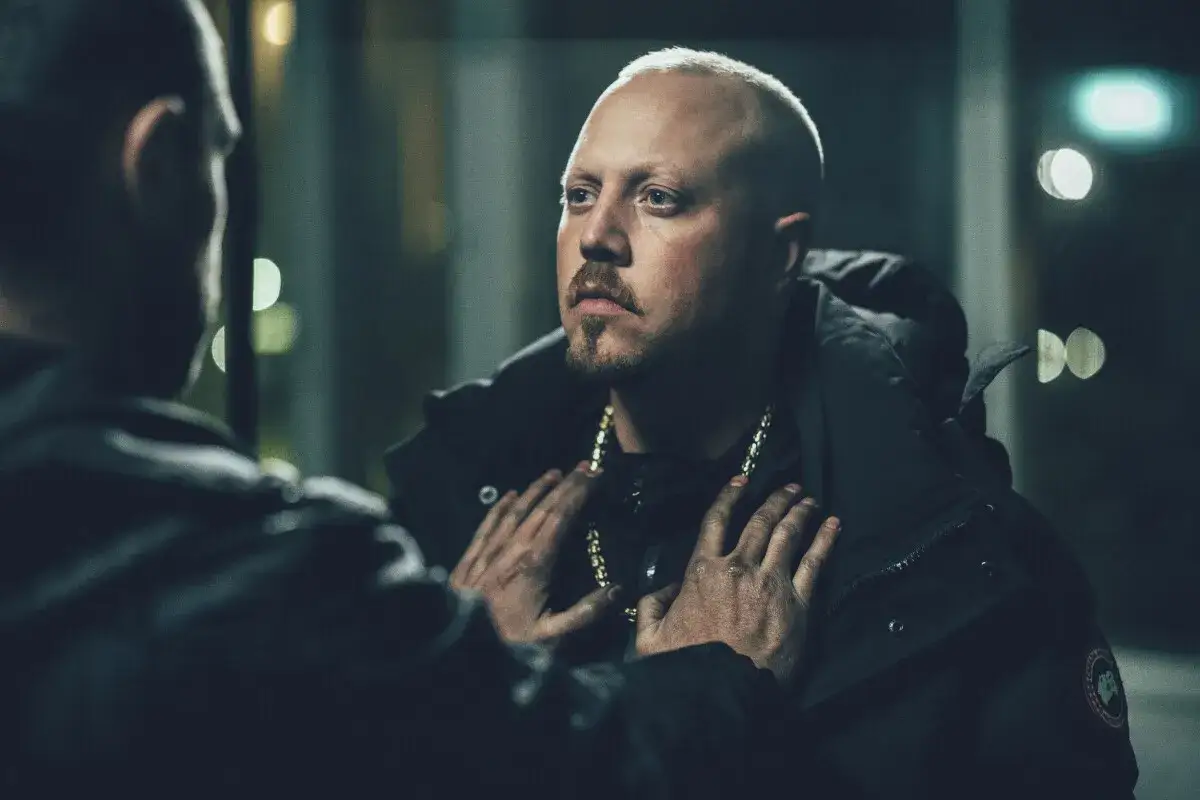 En intensiv scen från en sebbe stakset film. Två män möts i mörkret, en med guldkedja, den andra med händerna på hans bröst.
