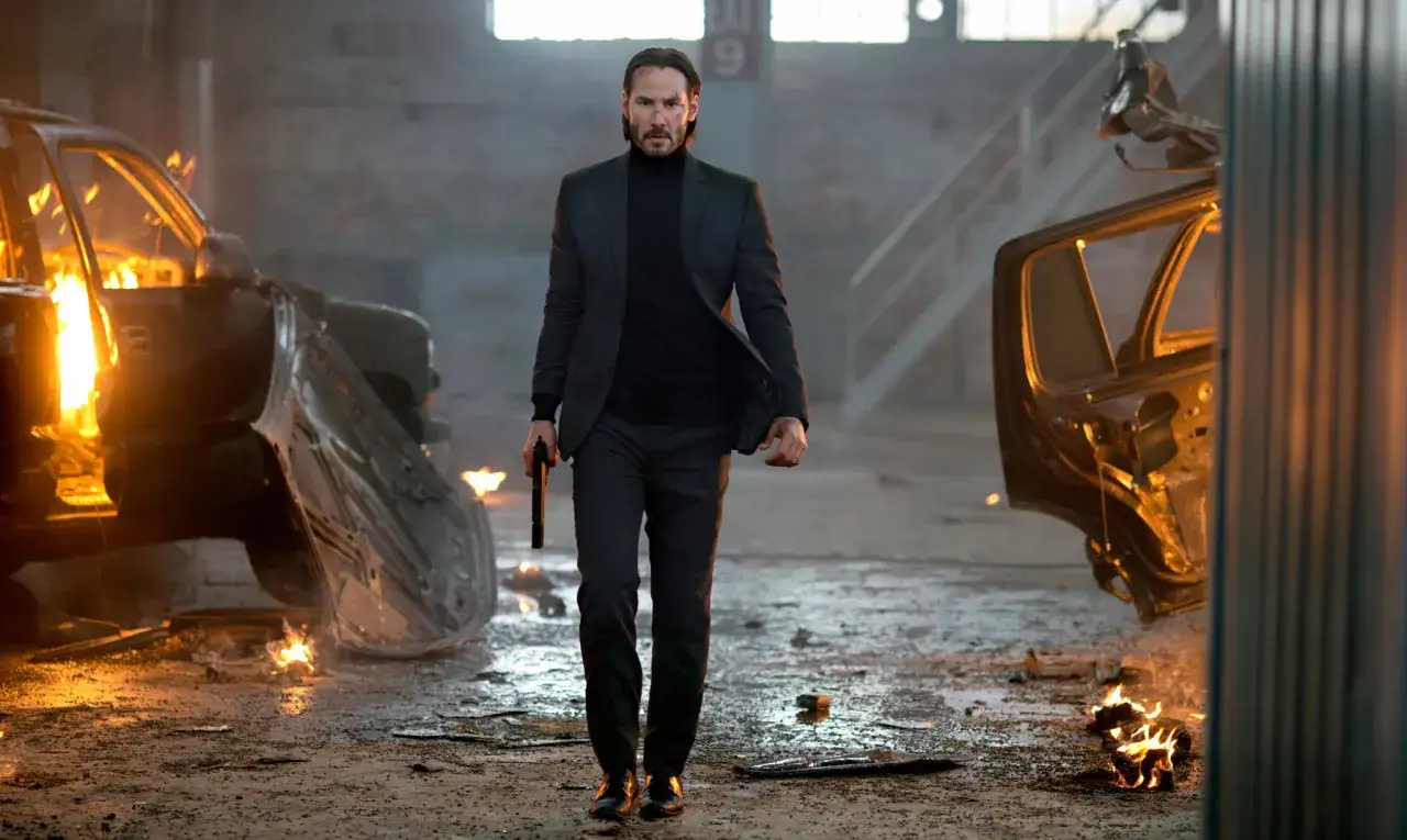 John Wick, med en pistol i handen, g&aring;r genom en lagerlokal med brinnande bilvrak.