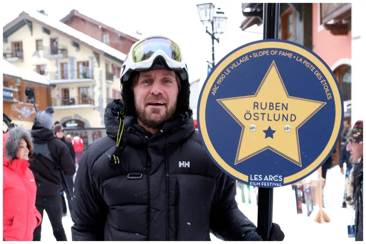 En skidåkare med skidglasögon och en HH-jacka står bredvid en skylt för